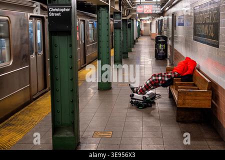 A person in a red jacket and plaid pants sits on a wooden bench in a subway station, resting their head on their arms while a small black device lies Stock Photo