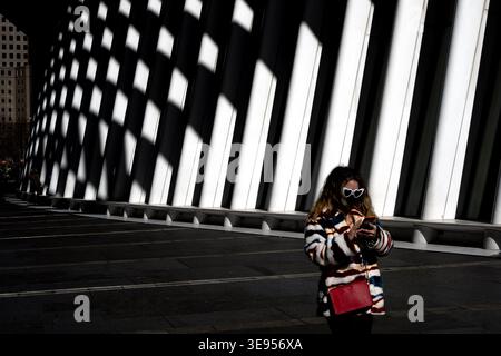 A person wearing sunglasses and a striped jacket walks along a modern bridge with strong diagonal shadows cast across the pavement. Stock Photo