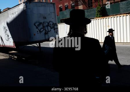 A person in a black suit and hat stands in the foreground while another person walks away in the background near a large white truck with graffiti. Stock Photo
