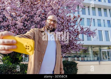 Smiling white man taking selfie on smartphone near blooming cherry tree in city Stock Photo
