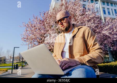 Confident man working outdoors on laptop in spring sunlight under cherry tree Stock Photo