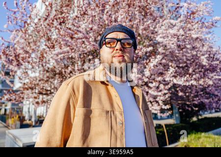 thoughtful bearded man in hat standing near blooming cherry tree Stock Photo