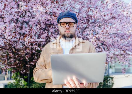 concentrated bearded man using laptop near blooming cherry tree Stock Photo