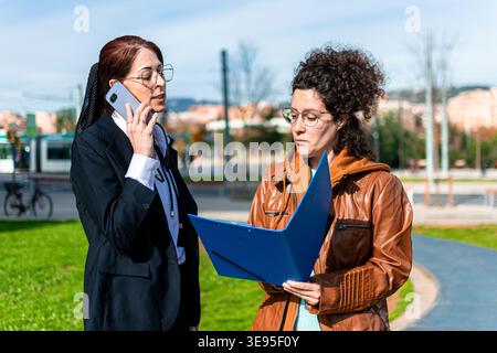 Businesswomen discussing project details and making a call outdoors Stock Photo