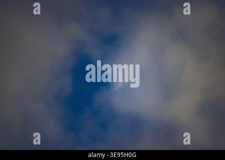 Crescent moon peeks through soft, drifting clouds. Dark blue sky behind. Clouds partially obscure moon. Serene night sky scene. Minimalist composition Stock Photo