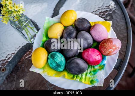 Dyed easter egg on colored napkin in plate, small vase with primrose on glass table. Stock Photo