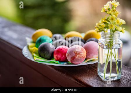 Dyed easter egg in plate, small vase with primrose on wooden railing, outdoor. Stock Photo