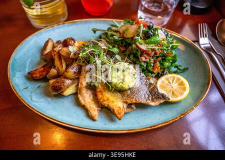 Roasted cod fish fillet with vegetable salad, baked potato and herb butter on blue plate, cutlery and drinks on restaurant table. Stock Photo