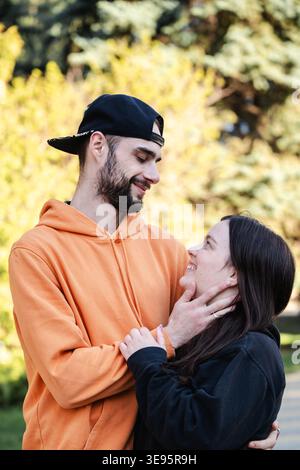 Young couple standing close together in park looking at each other and smiling while man touches woman face. Concept of love and romantic relationship Stock Photo