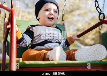 Infant child sitting on playground swing holding bars and smiling with joyful excited expression outdoors in sunlight. Concept of childhood emotion pl Stock Photo
