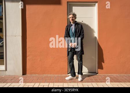 Full body man with mixed grey curly hair standing outdoors in front of an orange building wall and white door Stock Photo