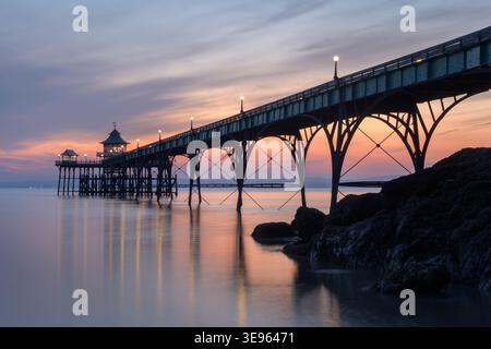 A neon mix of yellow, orange and pink colours at dusk illuminate the sky above Clevedon Pier, North Somerset. Stock Photo