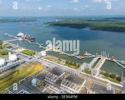 Aerial view of a bustling industrial port with ships docked, pipelines, and storage tanks. Clear skies over calm waters, surrounded by green hills. In Stock Photo