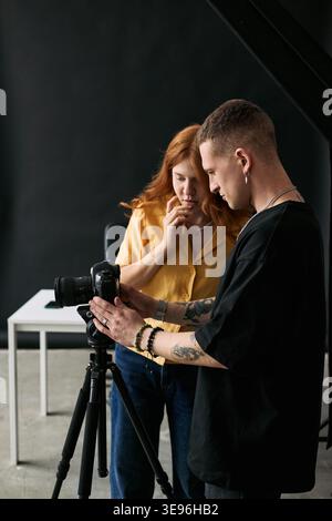 Vertical shot of young adult Caucasian man and woman reviewing camera footage on tripod during content production. Supporting blogging, livestream marketing, creator education, vertical Stock Photo
