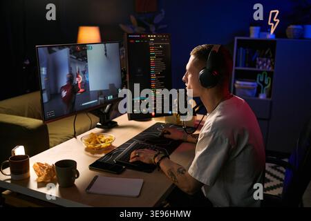 Young adult Caucasian man streaming gameplay on dual monitors, wearing headset and speaking into microphone. Useful for gaming content, live streaming, creator marketing, medium shot Stock Photo