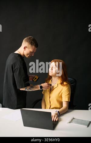 Young Caucasian woman sitting at laptop while man attaching microphone for livestream recording. Useful for blogging, content creation, influencer marketing, vertical Stock Photo