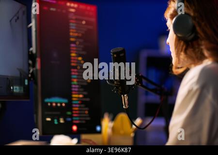 Young adult woman streaming live content with microphone, headphones, computer monitor, engaging online audience and creator economy workflow. Useful for blogging, livestream marketing, digital media Stock Photo