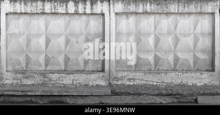 Weathered concrete fence featuring geometric relief panels and repeating textured pattern in monochrome. Urban architectural facade shows stains, deca Stock Photo