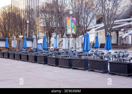 Outdoor seating area with rows of closed umbrellas and empty tables in urban park. Cold season city scene. Stock Photo