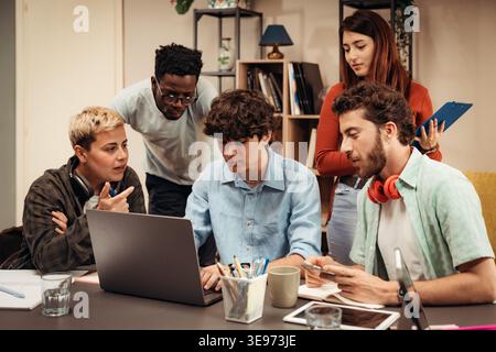 Diverse group of young professionals collaborating around a laptop during a project review meeting in a modern office. Teamwork, shared decision makin Stock Photo