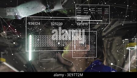Inspecting female tech in coveralls working on car undercarriage in bay, viewing LED light, holo UI Stock Photo