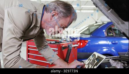 Leaning senior mechanic wearing coverall inspecting engine under hood at garage, using tablet Stock Photo