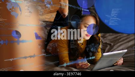 Reclining Asian woman with mustard top viewing tablet on dark-gray bed, holding phone, HUD overlays Stock Photo