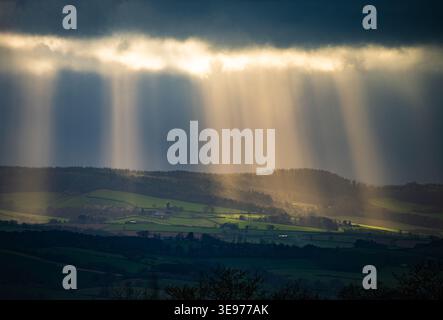 Dramatic sunbeams (crepuscular rays) breaking through dark storm clouds over the rolling green hills and patchwork fields of the Devon countryside. Stock Photo