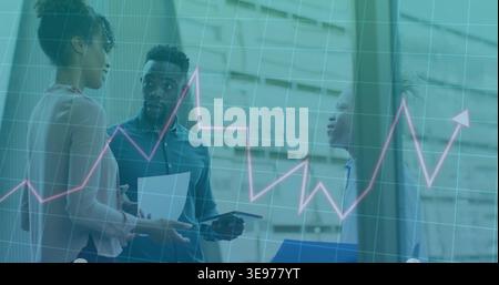 Analyzing line chart overlay, three colleagues in business wear discussing tablet, data in atrium Stock Photo