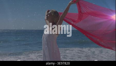 Adult woman in white sundress standing at beach holding sheer pink fabric, breeze lifting sun flare Stock Photo
