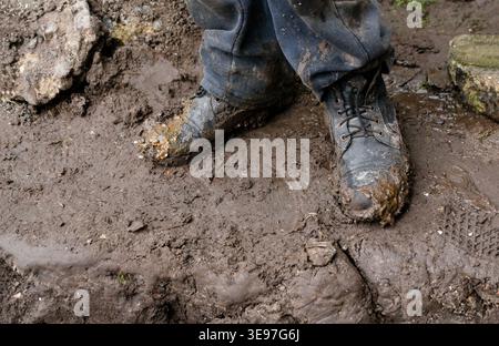 Close-up of black leather work boots covered in thick brown mud. Worker standing on a muddy construction site or rural field. Concept of hard manual Stock Photo