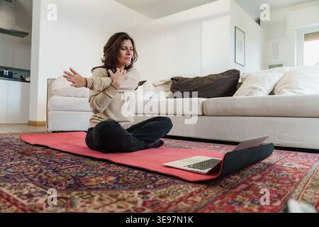 A woman is doing a stretching workout at home, sitting on a yoga mat with a laptop nearby. Stock Photo