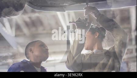 Working female technicians using socket wrench under raised car at shop, wearing coveralls goggles Stock Photo