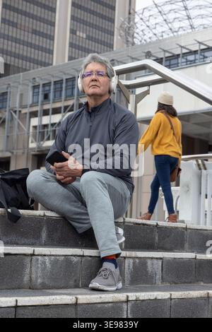 Senior man sitting on concrete steps in plaza with grey jacket headphones backpack holding phone Stock Photo