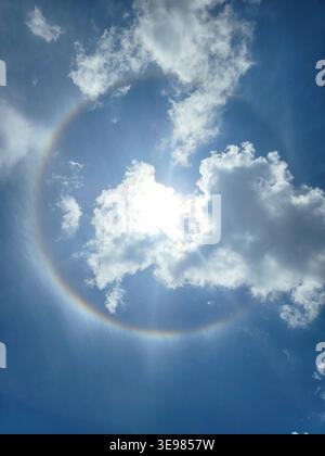 Sun shines bright through clouds. Circle of light surrounds the sun. Sky is vivid blue with white clouds. Natural phenomenon appears in sky. Perfect f Stock Photo