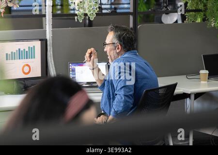 Colleagues watching man in blue shirt holding pen, working at office desk with laptop, monitor Stock Photo