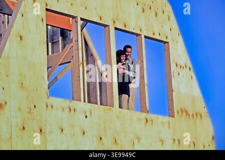 An African-American couple smiles and embraces while standing in a window of their new home under construction. Bright sunlight shines on them in the Stock Photo