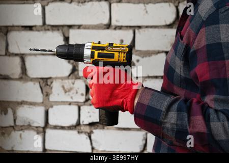 Worker in gloves holding cordless drill in front of brick wall. Construction, repair and home improvement concept. Stock Photo