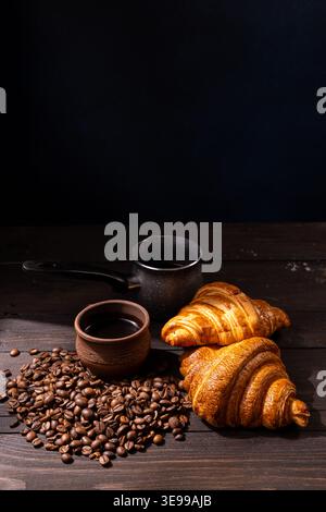 Freshly baked croissants, roasted coffee beans, a cup of brew, and a cezve arranged on a dark wooden table, creating a warm and rustic morning breakfa Stock Photo