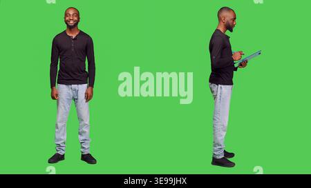 Confident man posing on camera and taking notes, writing important information using pen and clipboard papers. Casual male model standing over greenscreen bacground, leisure activity. Stock Photo