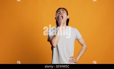 Drained person yawning and being tired in studio, falling asleep and feeling worn out. Exhausted young man being sleepy and carefree over orange background, having confidence on camera. Stock Photo