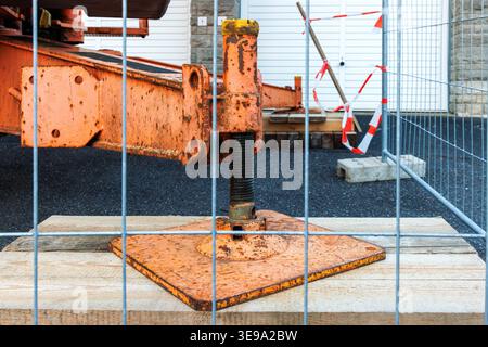 Close up of a rusty crane outrigger support foot on a metal base plate through a temporary safety fence at a roadwork construction site. Stock Photo
