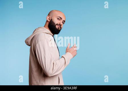 Smiling arab man inviting with hand and looking at camera with relaxed facial expression. Young bald bearded person asking to come over with arm gesture, posing for studio portrait Stock Photo