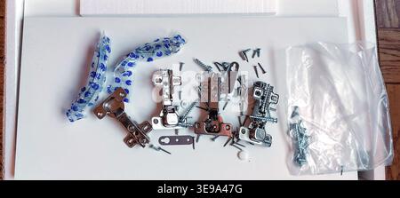 A top-down view of various cabinet hinges, screws, and metal handles laid out on a white furniture board, ready for assembly. DIY home improvement and Stock Photo
