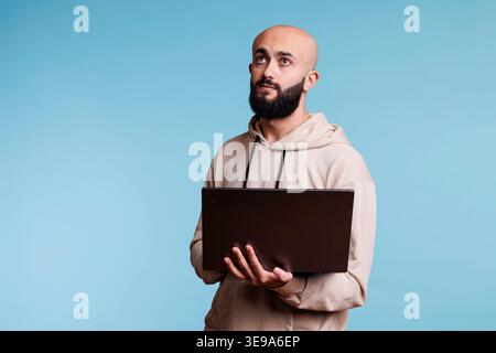 Thoughtful arab man holding laptop and looking upwards with pensive expression. Young person in doubt programming software application while standing with portable computer Stock Photo
