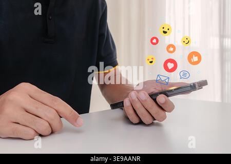 A man using a smartphone with social media emojis floating on the screen. Online communication. Stock Photo