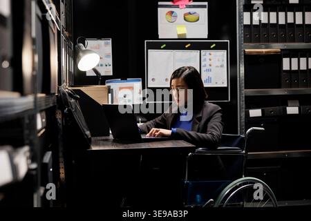 Professional businesswoman accountancy employee in wheelchair writing budget plan and accountancy strategy on laptop. Asian woman in repository filled with document folders and flowcharts Stock Photo