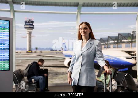 Woman passenger portrait, carrying luggage for business trip, at boarding gate, walking around the airport terminal before departure. Female traveler checking flights information on screens. Stock Photo