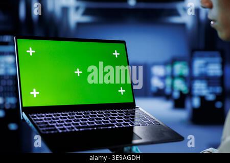 IT consultant with chroma key laptop between server hub clusters providing processing resources for businesses worldwide. Worker uses isolated screen device to fix data center mainframes Stock Photo