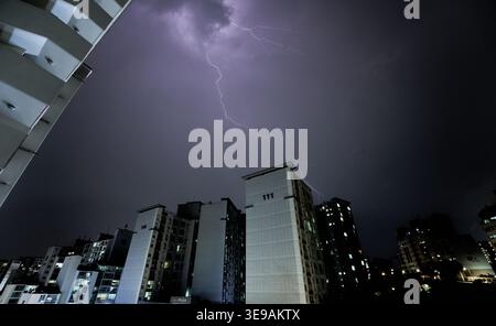 A powerful lightning bolt strikes over a modern apartment complex at night. The purple storm clouds and silhouetted buildings create a dramatic scene. Stock Photo